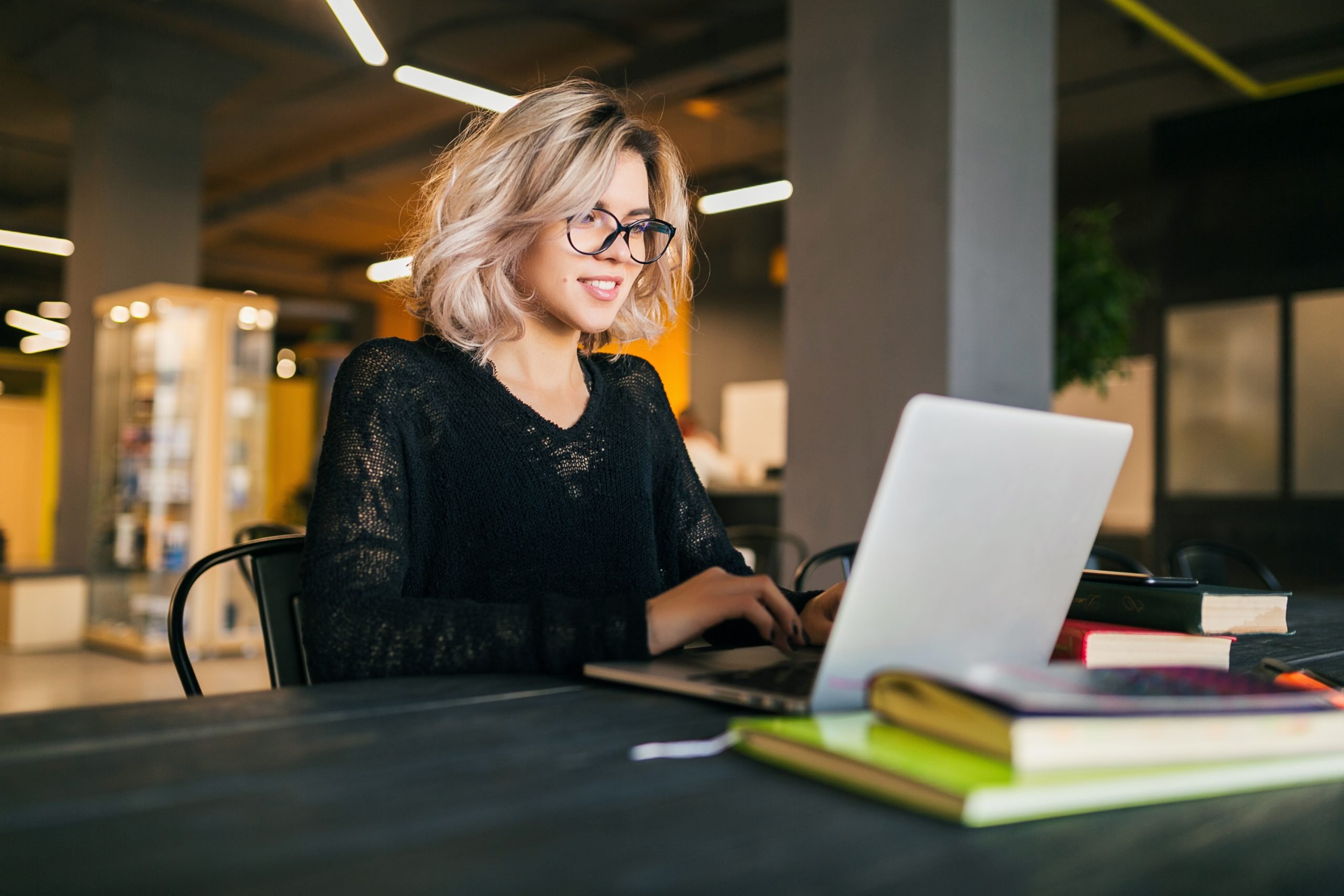 portrait of young pretty smiling woman sitting at table in black shirt working on laptop in co working office wearing glasses scaled